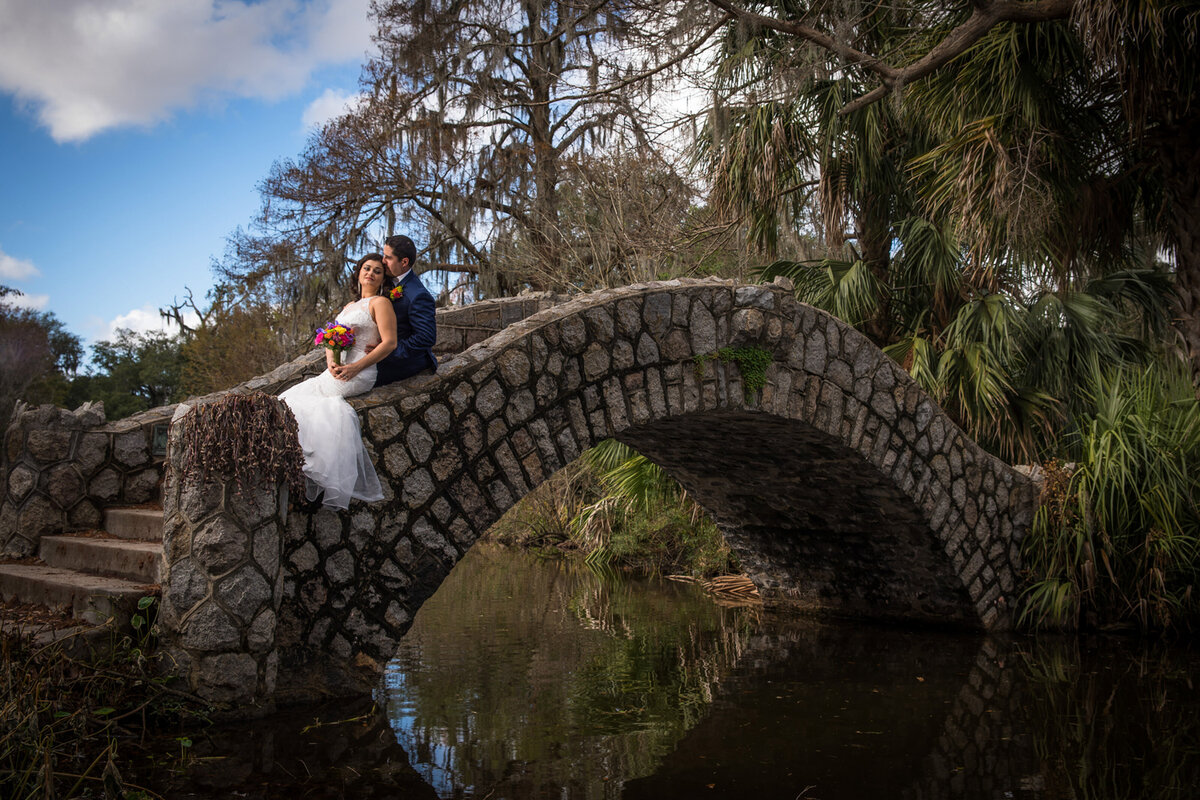Groom embraces his bride on stone bridge over park pond