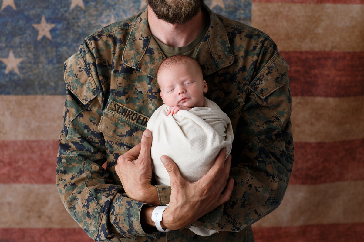 Dad dressed in military fatigues holding  his infant son photographed on an american flag background