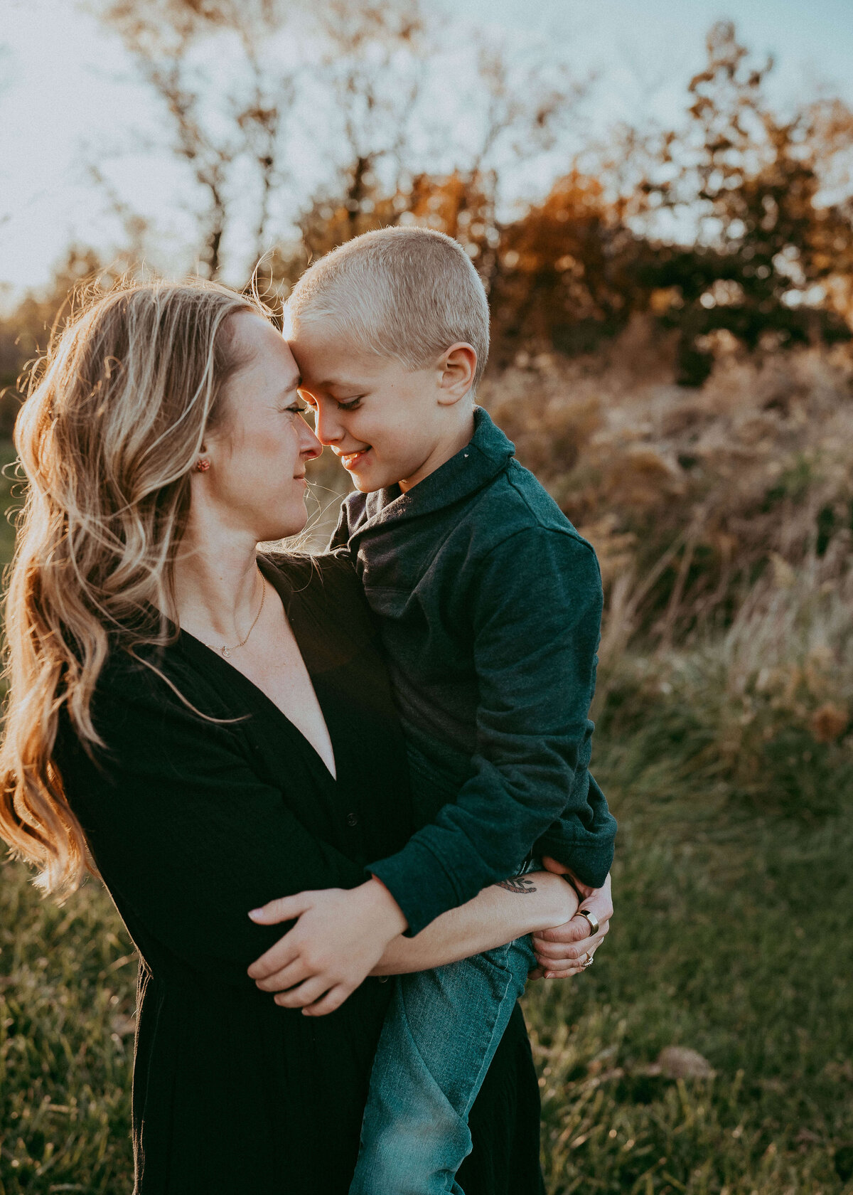A gorgeous, fine art image of a mothers silhouette being back, lit by the sun as she holds her young son, while they face each other with their foreheads touching.