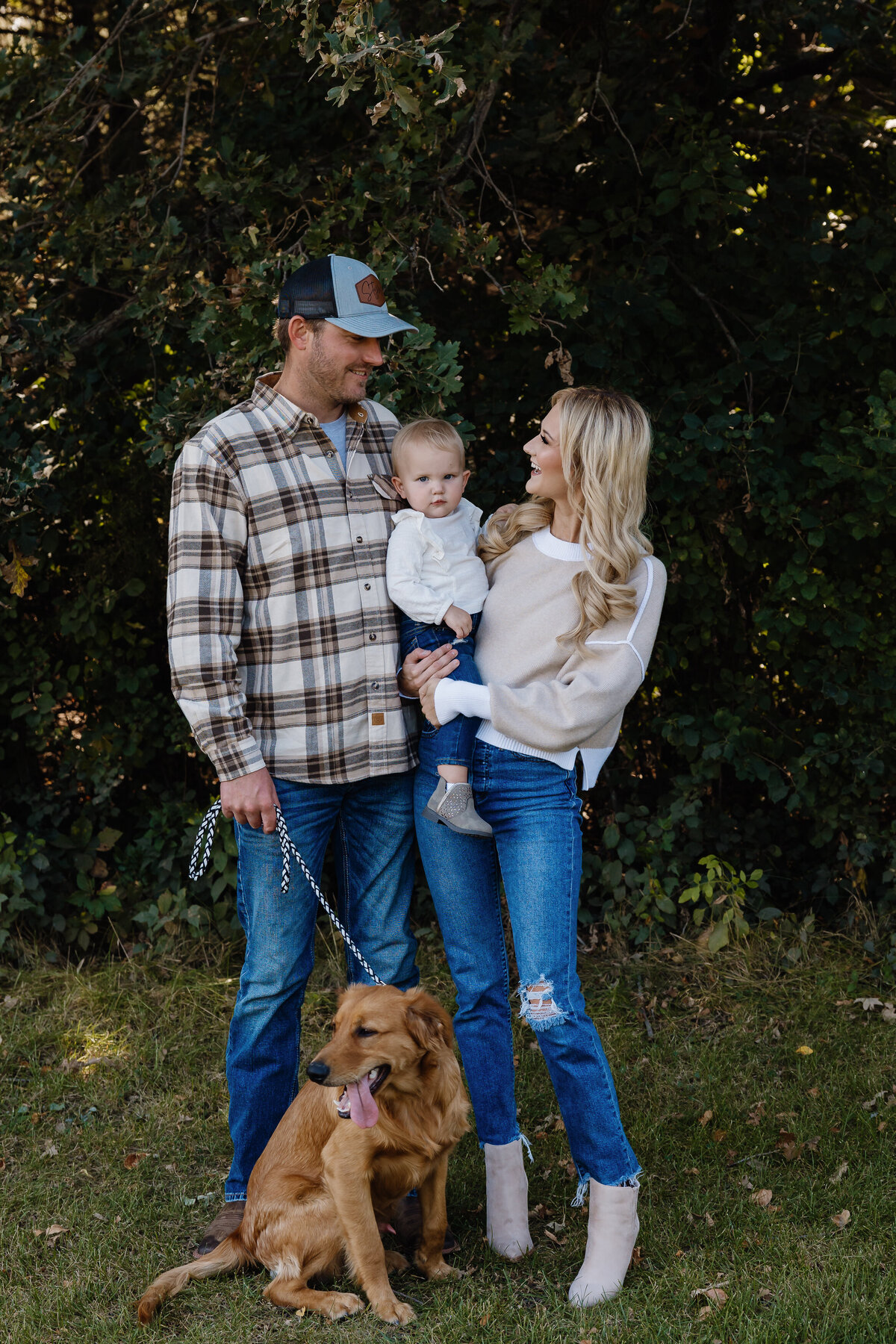 Two parents smiling while one holds a child and the other holds the leash of a dog.