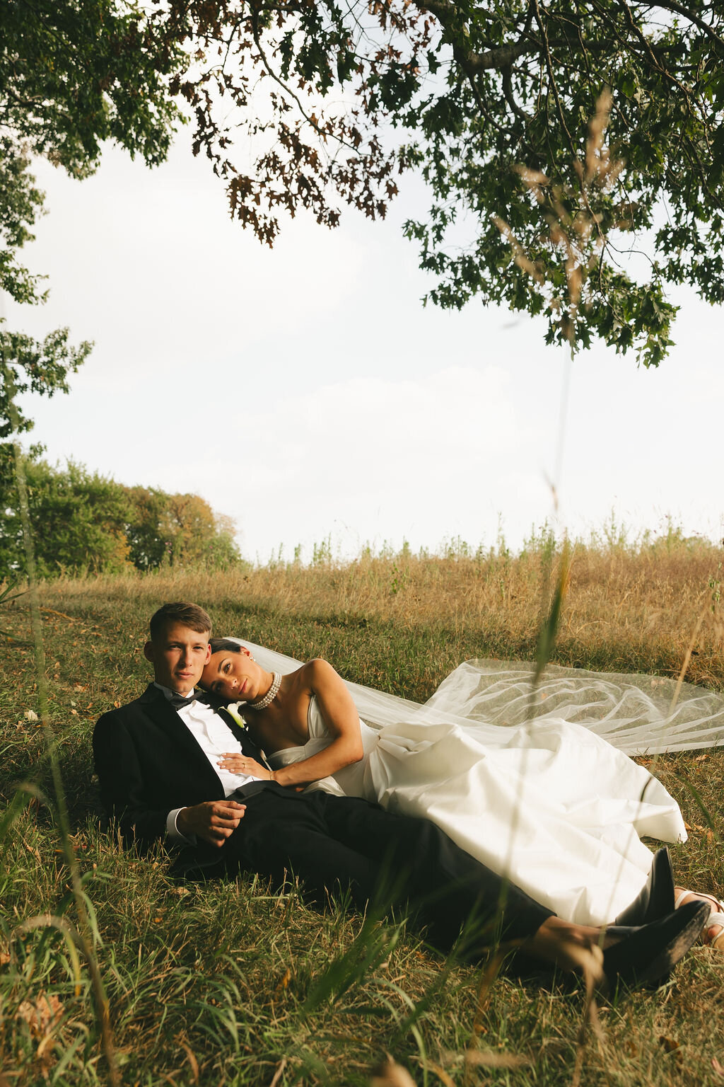 Bride and groom facing the camera holding hands while looking apart from eachother. They are standing in front of deep green trees