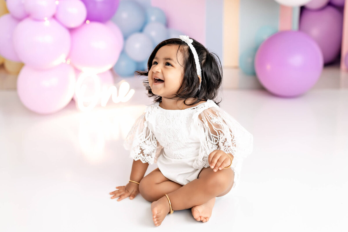 A joyful toddler wearing a white outfit and headband sits on a glossy floor, surrounded by pastel balloons in pink, purple, and blue. The child smiles while looking slightly upwards.