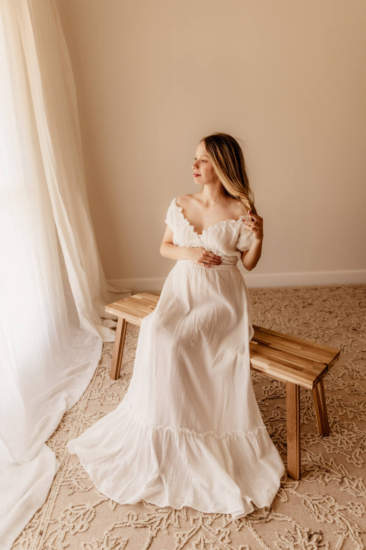 Woman in her third trimester sitting on a bench and twirling her hair wile looking out the window for an OKC maternity photoshoot.