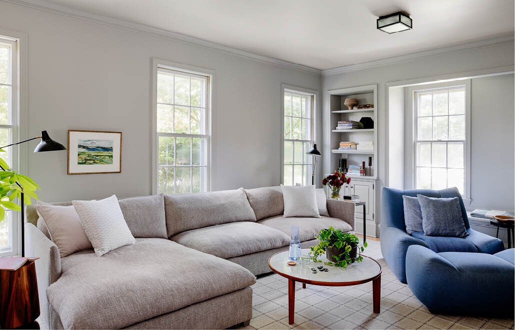 Experience the bright family room in a historic Hingham home, featuring a light grey sectional with chaise, denim-upholstered lounge chair and ottoman, and a marble and walnut coffee table.