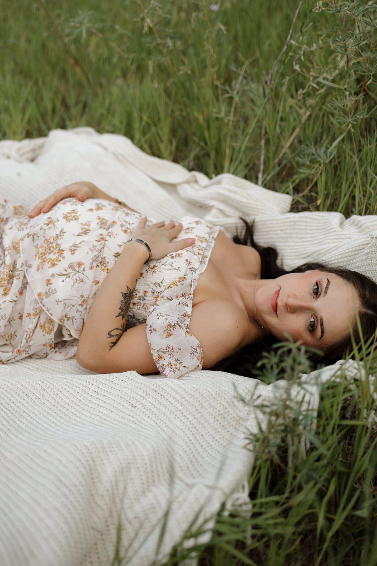 Expectant mom laying on white blanket in meadow in boulder colorado holding belly with eyes opened while smiling during maternity session with alexis adkins photography