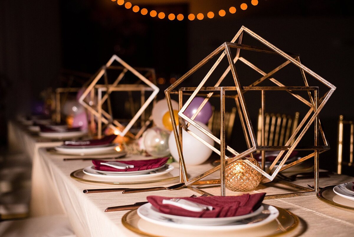 Long table with gold table cloth set with gold chargers, marble plates, burgundy fancy folded napkins and marble tile seating cards. Large gold geometric centerpieces interspersed with small clusters of balloons and gold votive candles.