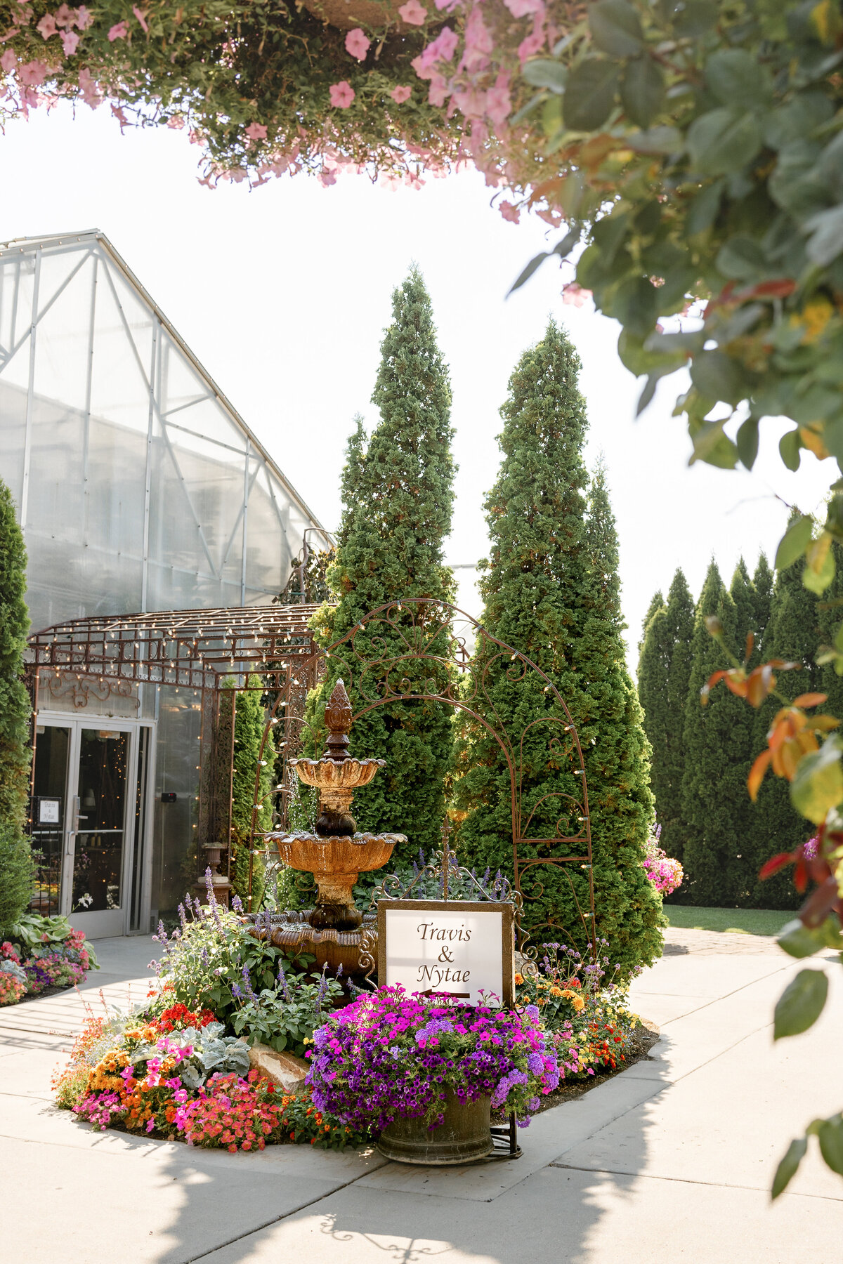A beautifully landscaped entrance to Le Jardin in Sandy, Utah, featuring a grand stone fountain, vibrant flower arrangements, and a sign welcoming guests to the wedding of Travis & Nytae.
