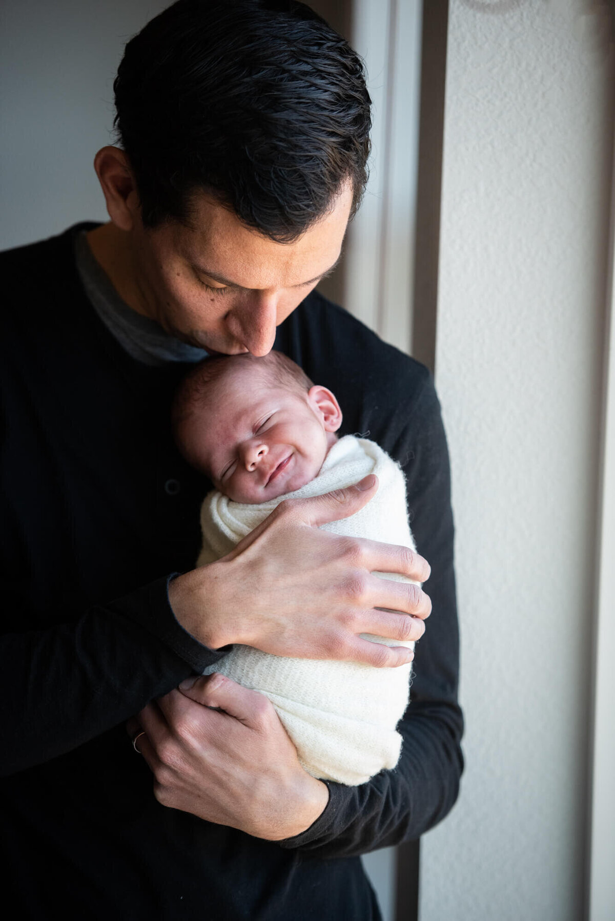 A newborn baby smiles while being kissed by dad as he stands in a window