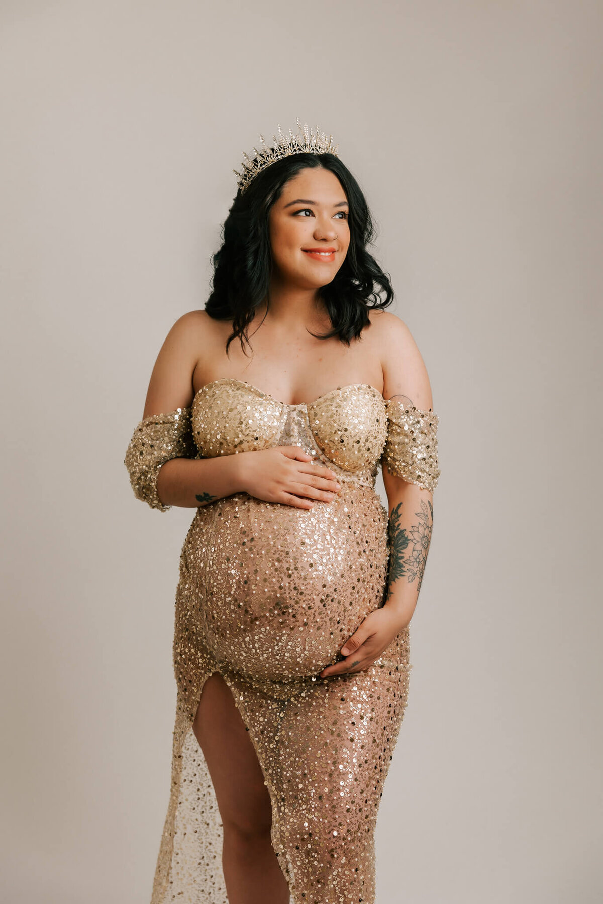 beautiful portrait of woman wearing gold sequin gown and crown. She has her hands on her pregnant belly.