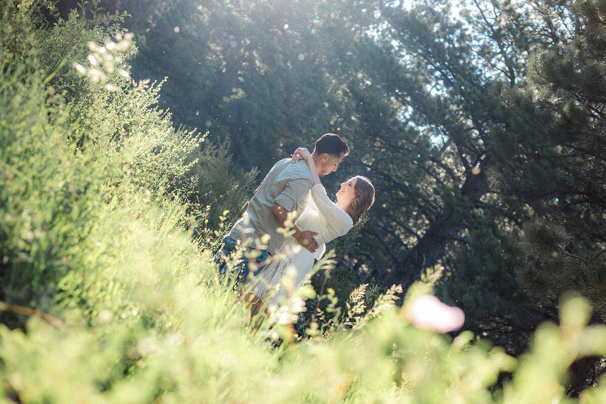 The couple embraces amidst sunlit green bushes and tall trees, creating a dreamy, romantic setting.