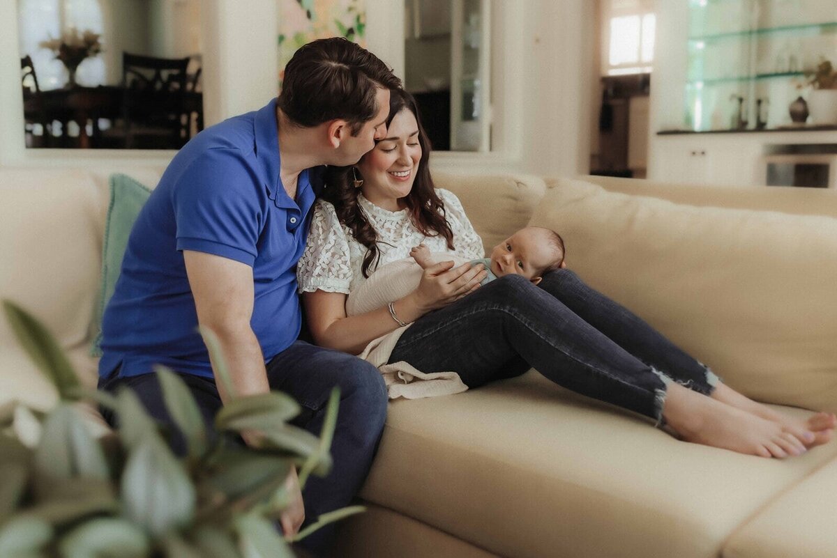 A couple sits on a couch; the man kisses the woman's forehead as she holds a baby. The living room has a dining area and a plant in the foreground.