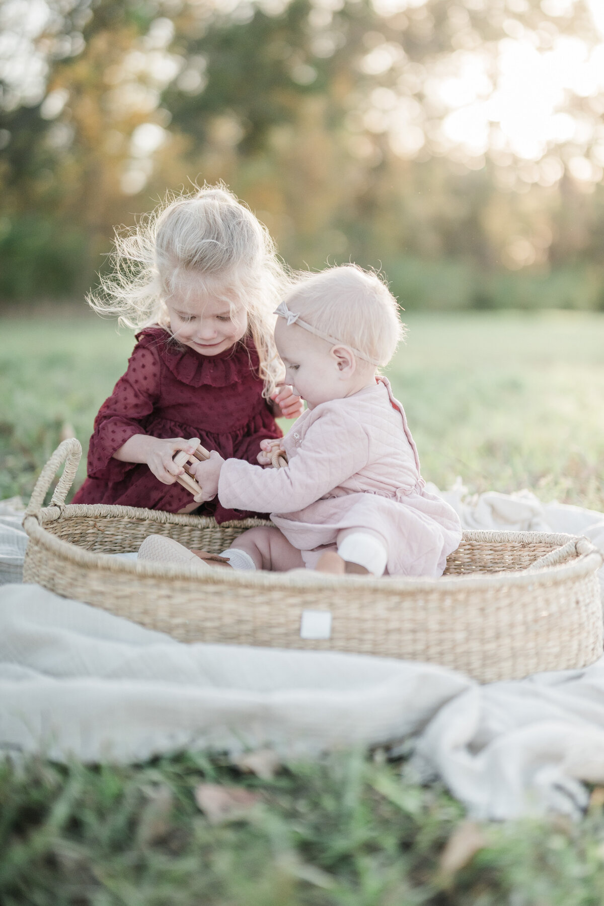 Older sister gently playing with baby sister sitting in a woven basket outdoors during a Kansas City milestone session.