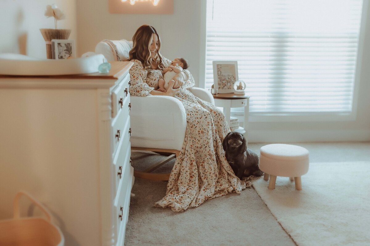 In a softly lit room, a woman gently cradles her baby in a rocking chair, embodying the serene and tender moments captured by a Macon newborn photographer. Nearby, the loyal family dog rests on the floor, completing this peaceful family portrait.