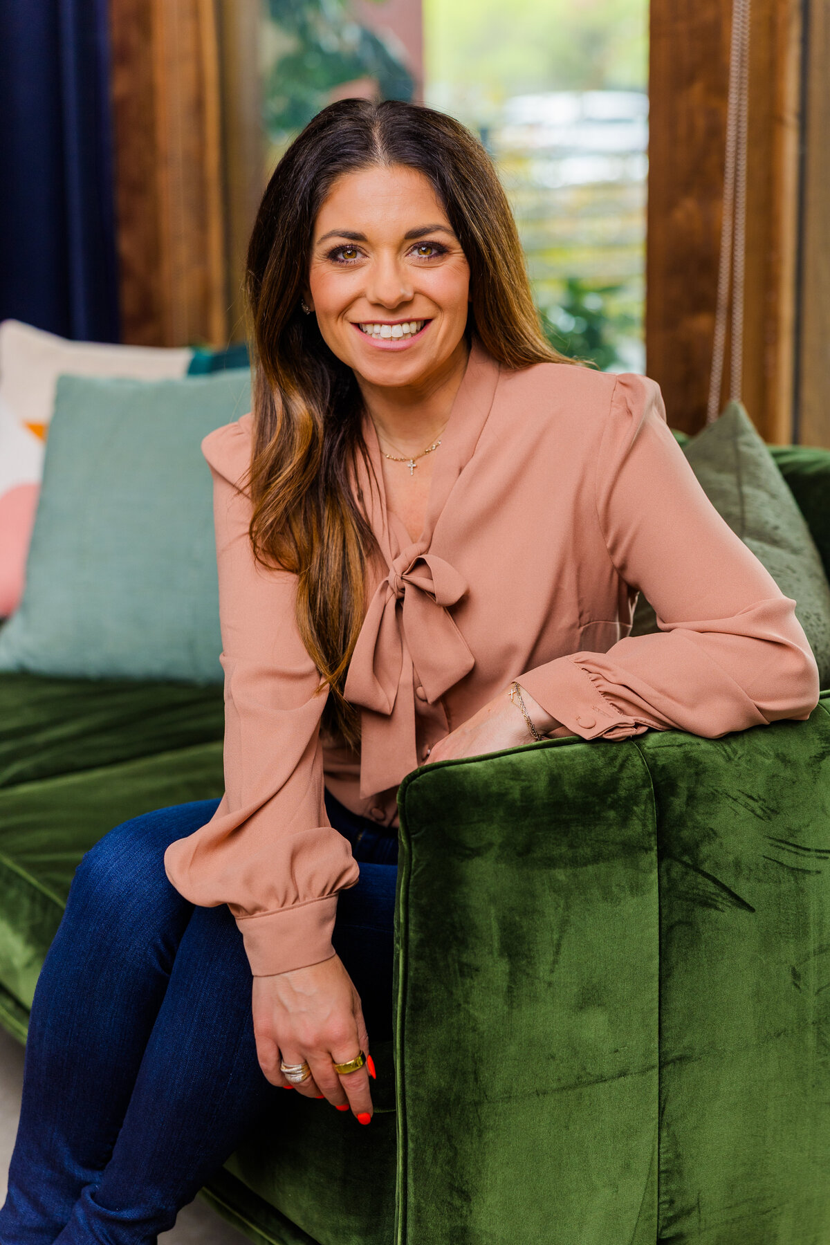 woman wearing a light pink top over a pair of jeans sitting on a green sofa, one arm resting on the armrest during portraits in studio