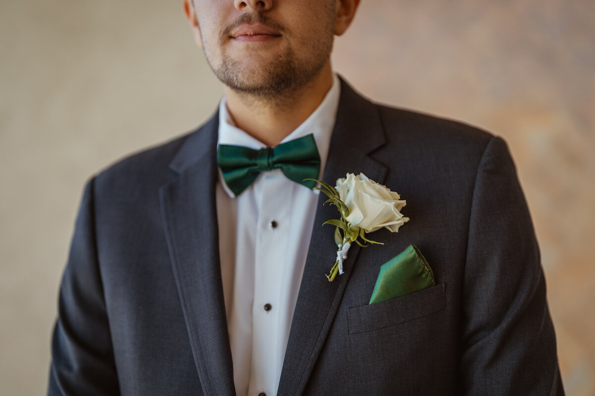 Close-up of a groom's wedding attire featuring a green bow tie, white rose boutonniere, and matching pocket square, taken during a DFW wedding. The image highlights the elegant details of the groom's ensemble.