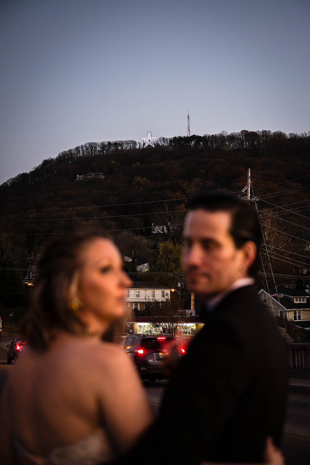 A bride and groom wrap their arms around one another and look in opposite directions with the Mill Mountain Star in the background. The couple in the foreground are purposefully blurred out to focus on the now illuminated Roanoke Star atop Mill Mountain at sunset.