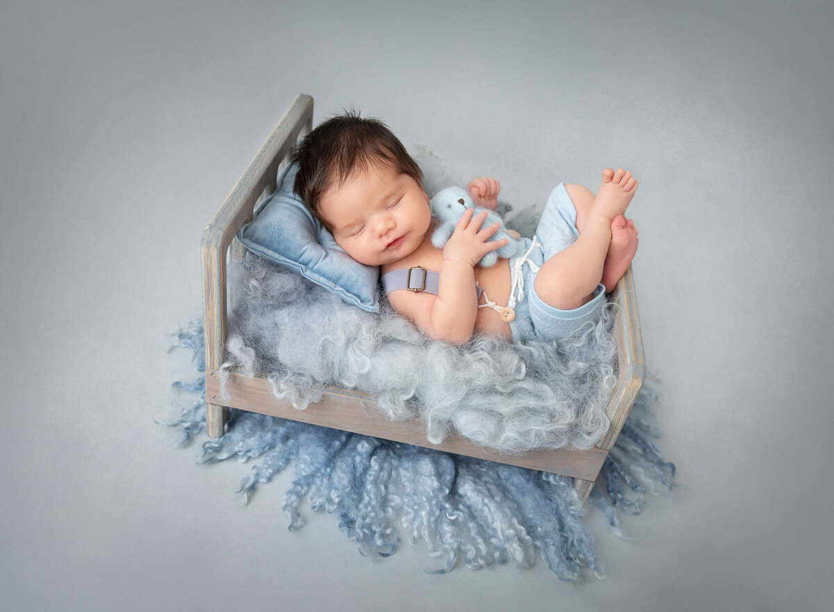 Newborn baby  boy grey and blue set up baby boy smiling wearing light blue shorts and holding tiny white teddy bear