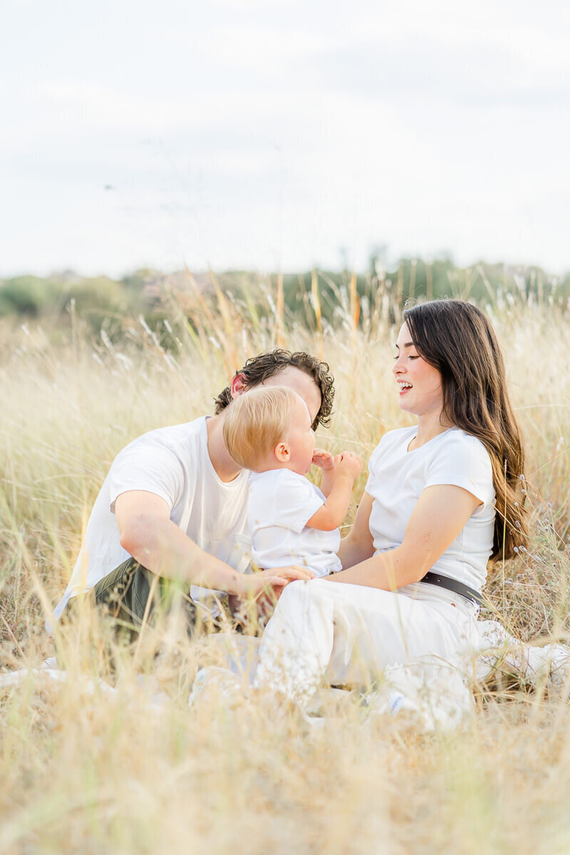a mother and father sit with their toddler boy in a field of tall grass. They play candidly while being captured by their Austin family photographer