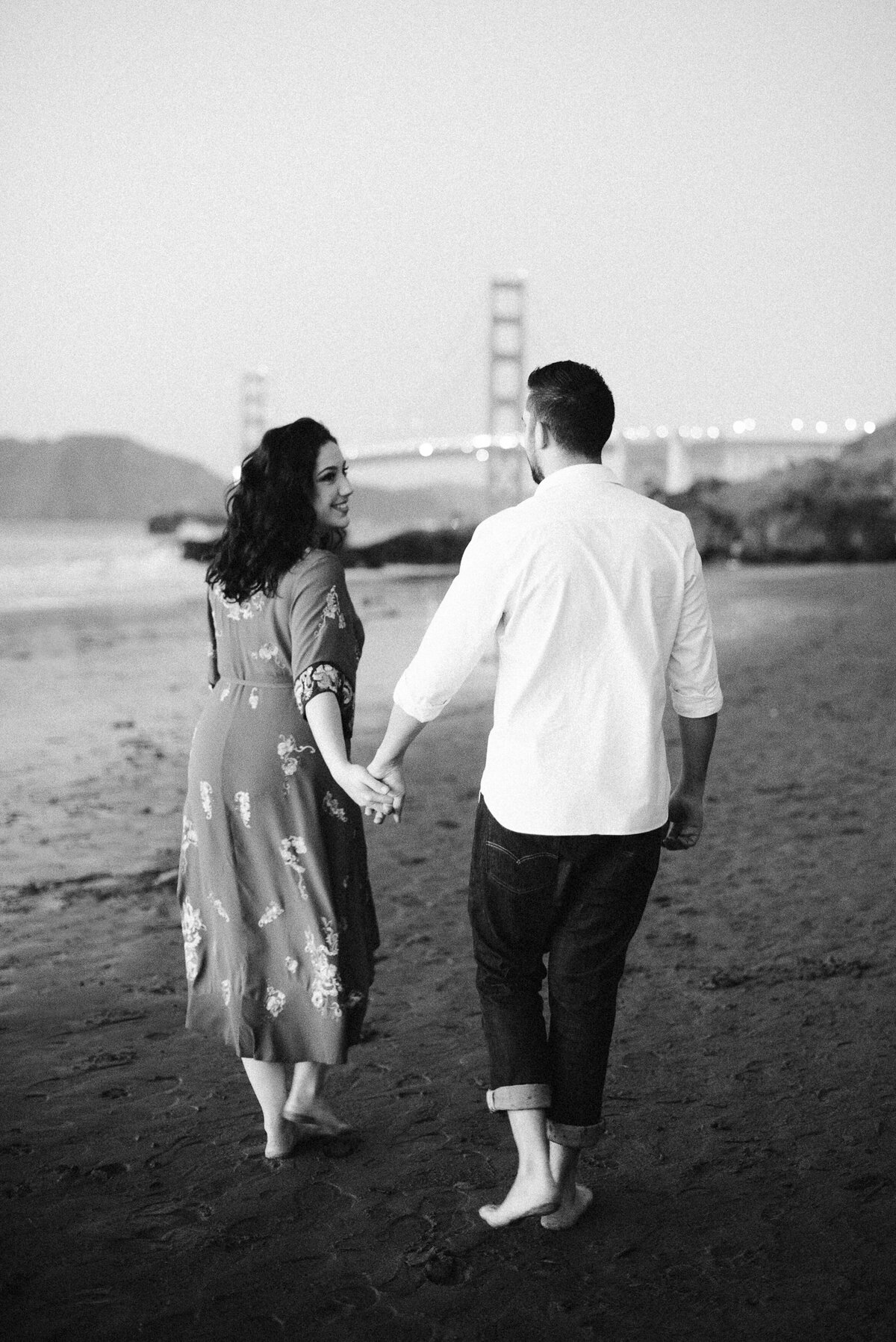 Classic black and white engagement photography at Baker Beach, San Francisco. Timeless candid moment with couple walking barefoot along the shore, featuring the iconic Golden Gate Bridge in the background. Floral midi dress and casual white shirt styling. Fine art film photography capturing authentic San Francisco landmarks.
