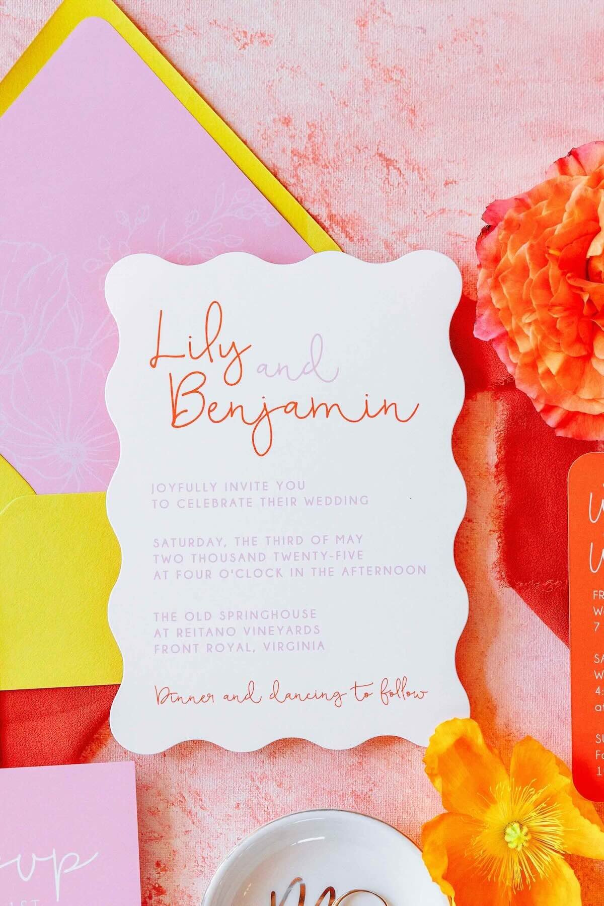 Scalloped wedding invitation flatlay in pink and orange, accented with bright flowers and colorful paper layers.