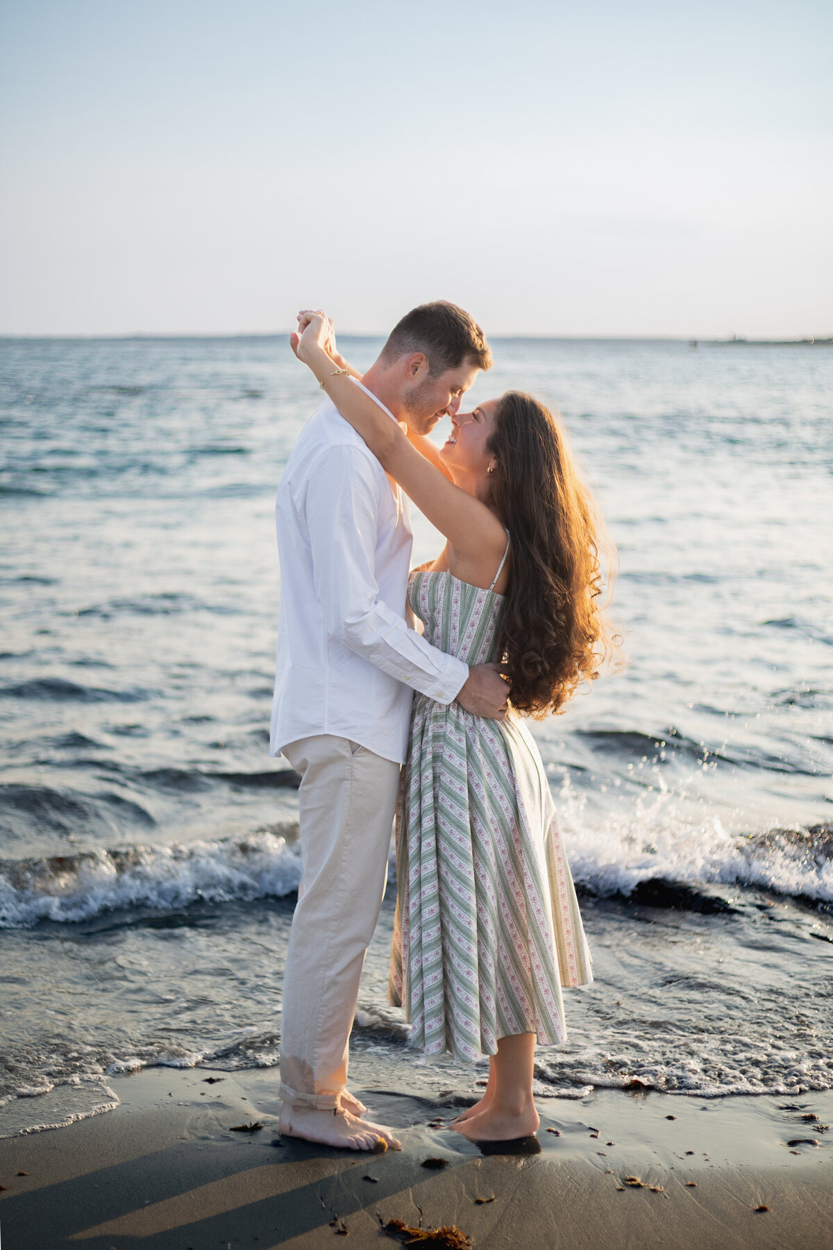 Collins Beach Newport RI | Kelsey Sheehan Photography Timeless Rhode Island Weddings | A couple embraces near the ocean, waves gently lapping at their feet. The man in a white shirt and the woman in a striped dress gaze at each other lovingly.