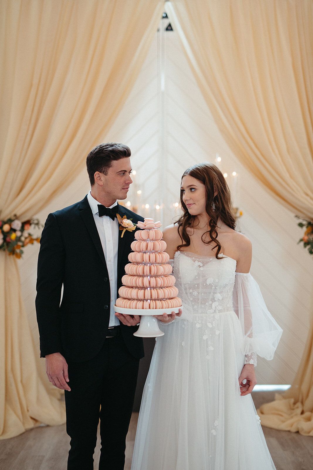 Bride and groom wearing a white wedding gown and black tuxedo holding a cake stand filled with blush macaroons.