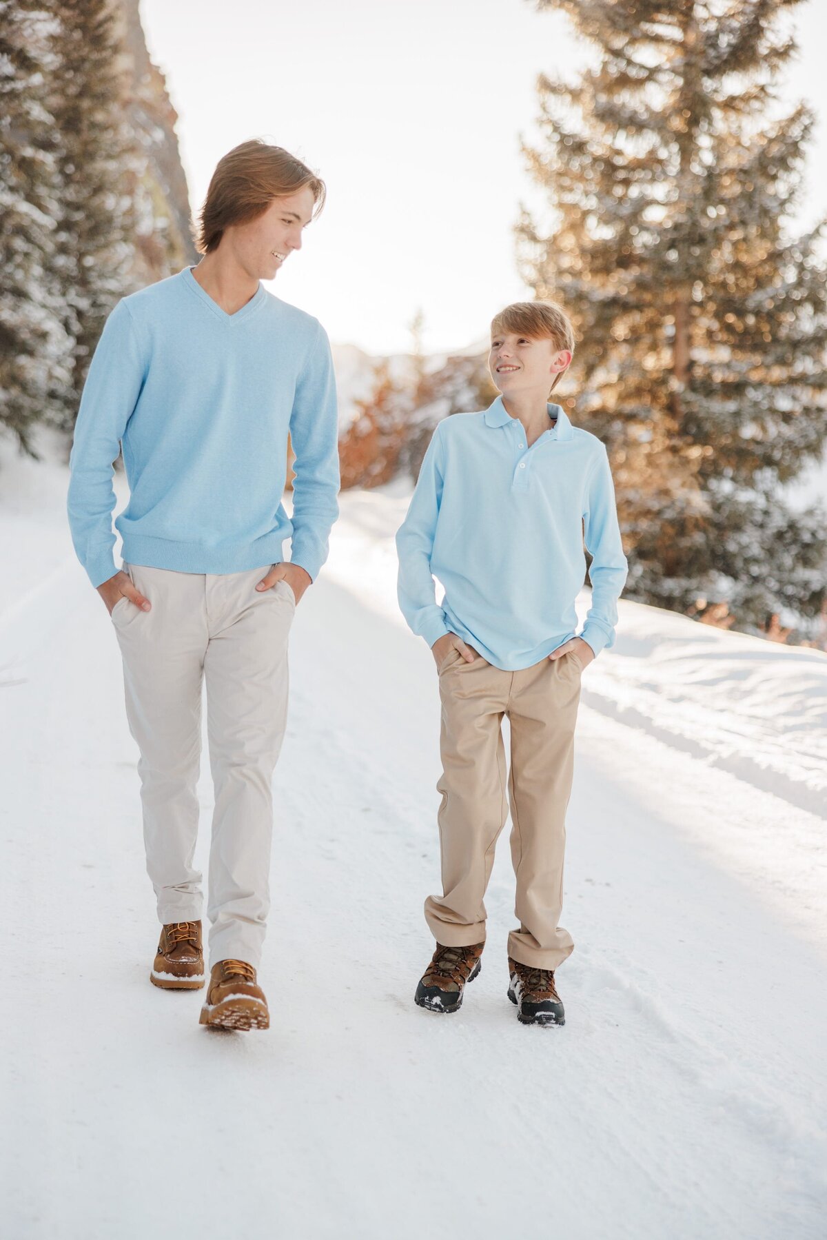 Smiling teen brothers in blue shirts and khakis walk together in a snow covered trail