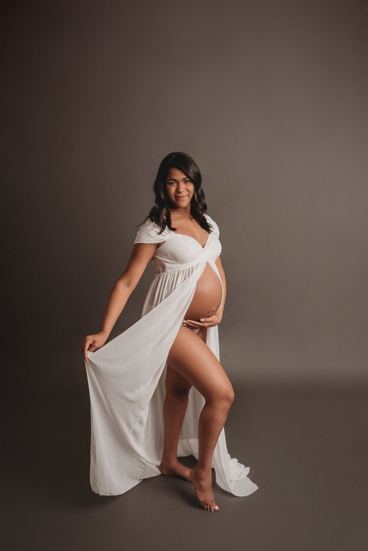 Pregnant woman posing for maternity pictures in a white sheer dress and grey backdrop.