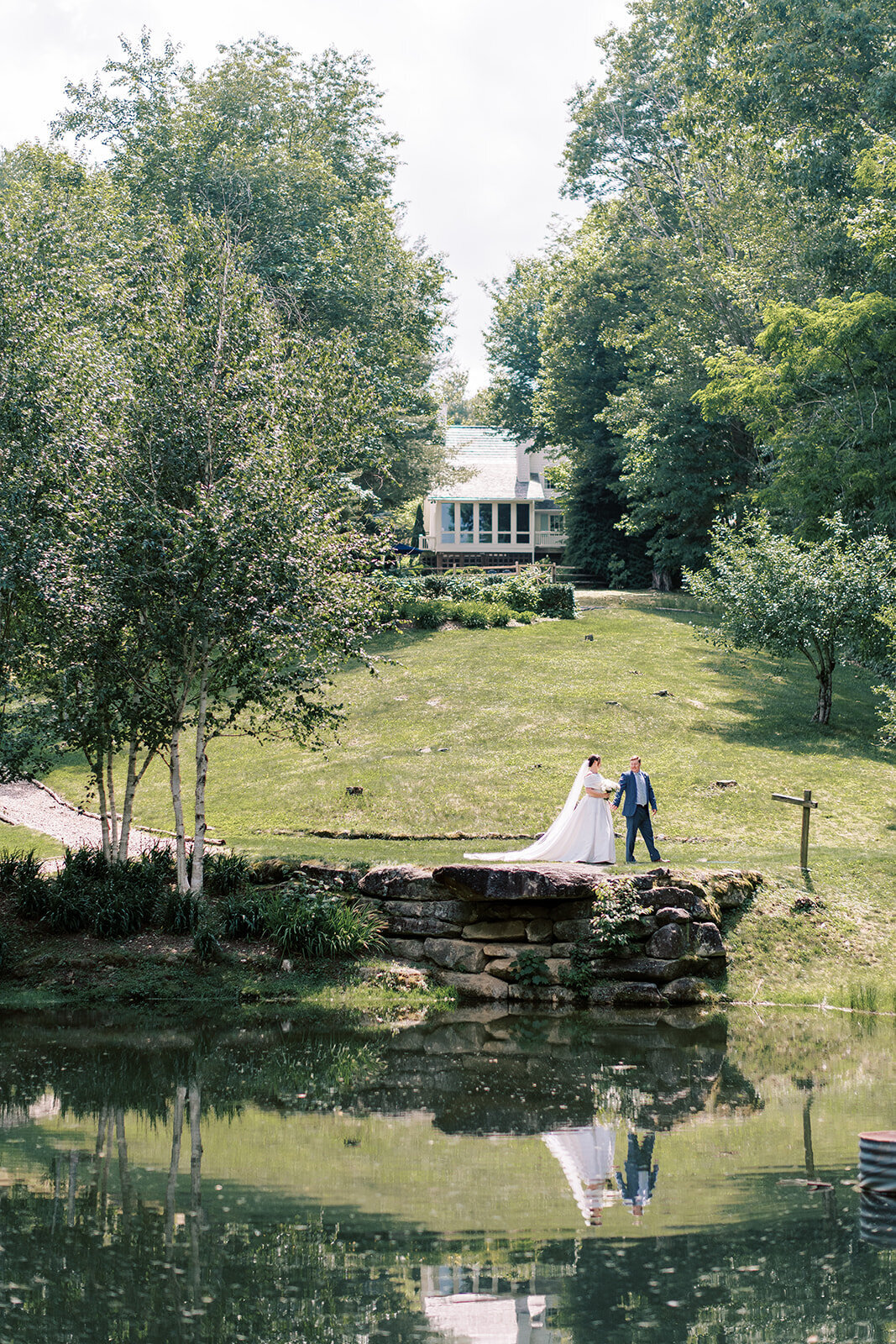 Bride and groom walk across a stone bridge with their reflection in the water at a private estate mountain wedding in Cashiers NC.