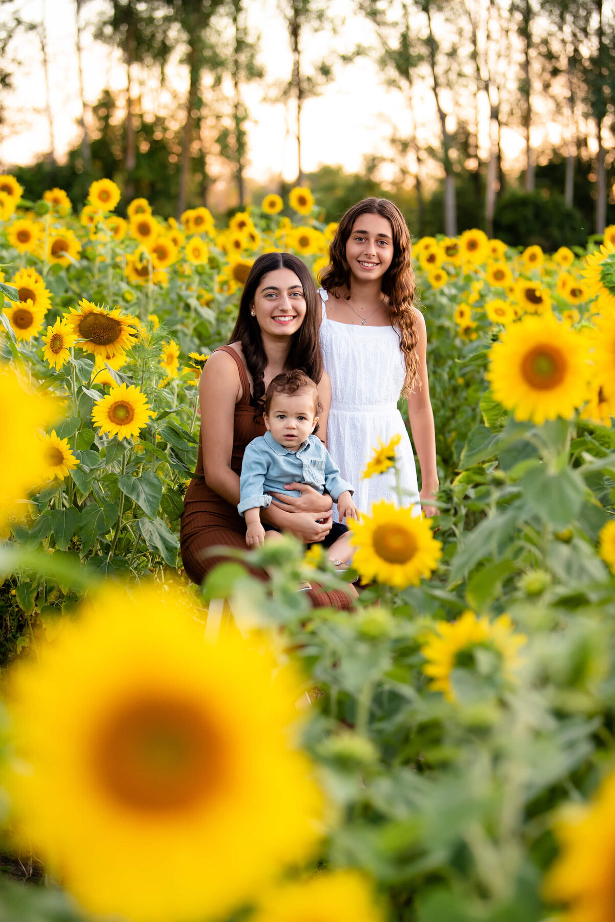 two teenage girls with their baby brother in a sunflower field for their summer family photography session