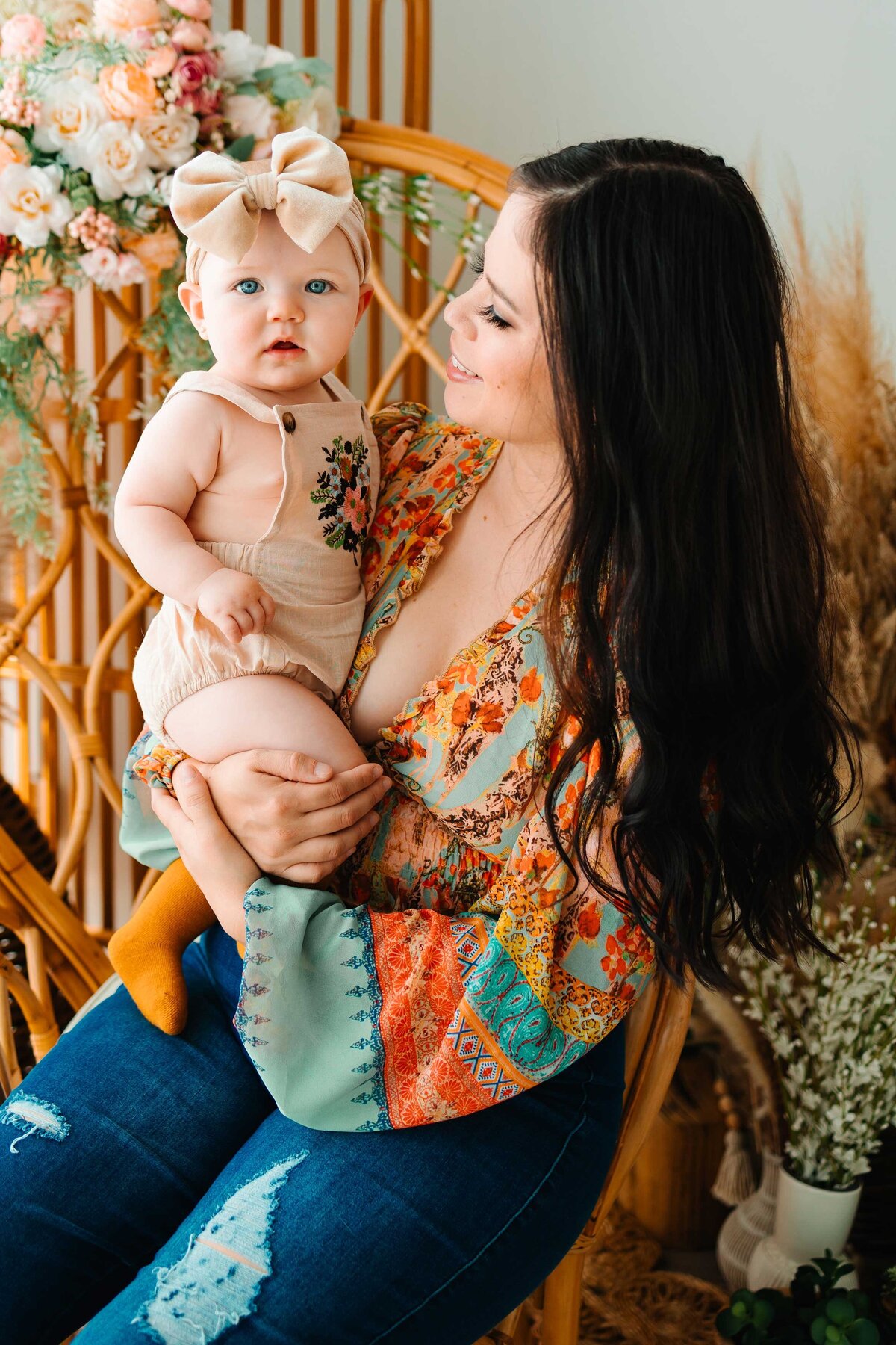 A joyful mom carries her little baby, both dressed in vibrant outfits. The scene, captured by a top Albuquerque photographer, features a charming backdrop with a chair and decorative faux flowers, showcasing their happy and loving moment.