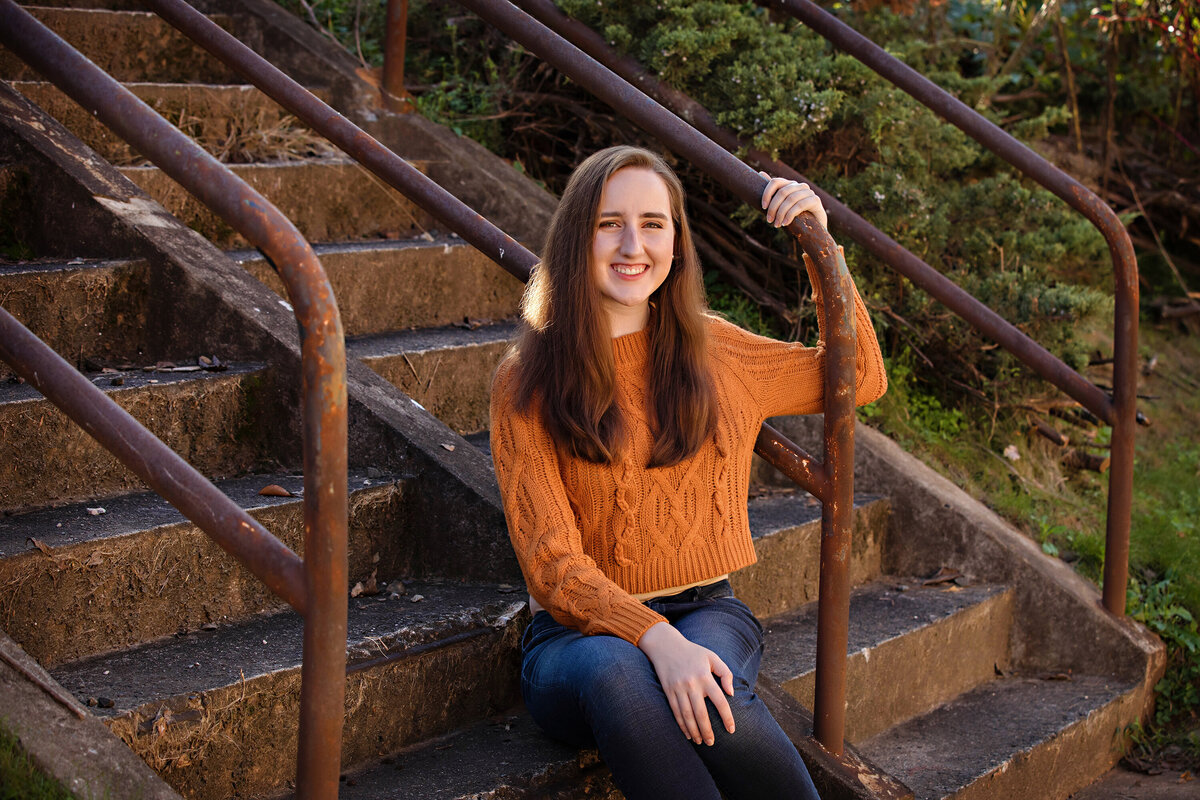 Beautiful HS senior in orange sweater sitting on concrete staircase with rust‑colored railings and greenery at Camp North End Charlotte NC