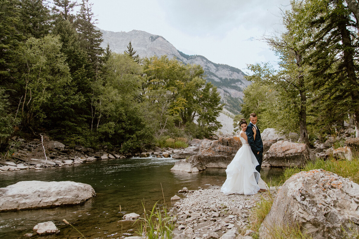 Bride and groom embraced along the waters edge in Crowsnest Pass, captured by Love and be Loved Photography, authentic and natural wedding photographer and videographer in Lethbridge, Alberta. Featured on the Bronte Bride Vendor Guide.