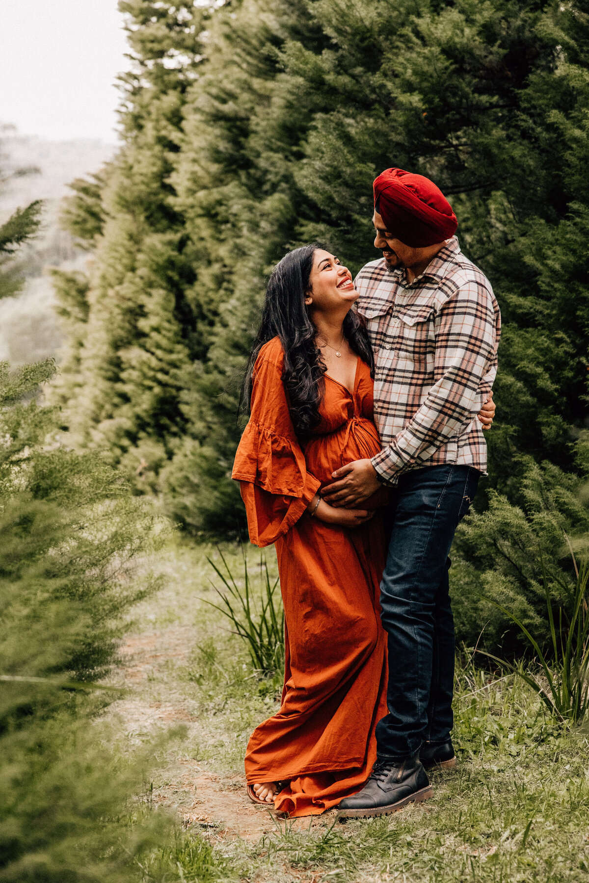 Expectant couple cuddle and look at each other while standing amongst trees in Melbourne Dandenong Ranges