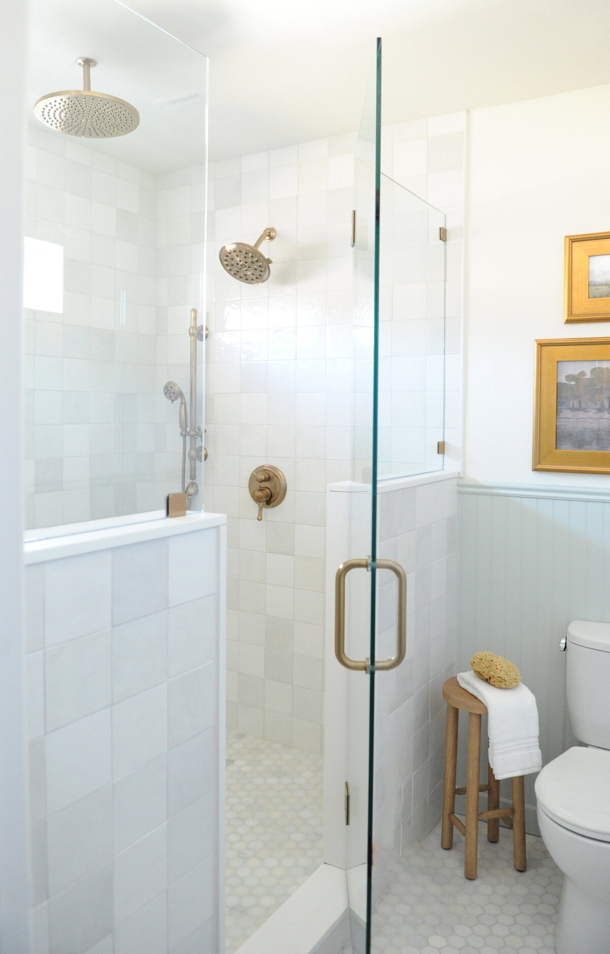Glass shower with brass plumbing fixtures and rain shower head, wooden stool, and gold framed landscape paintings