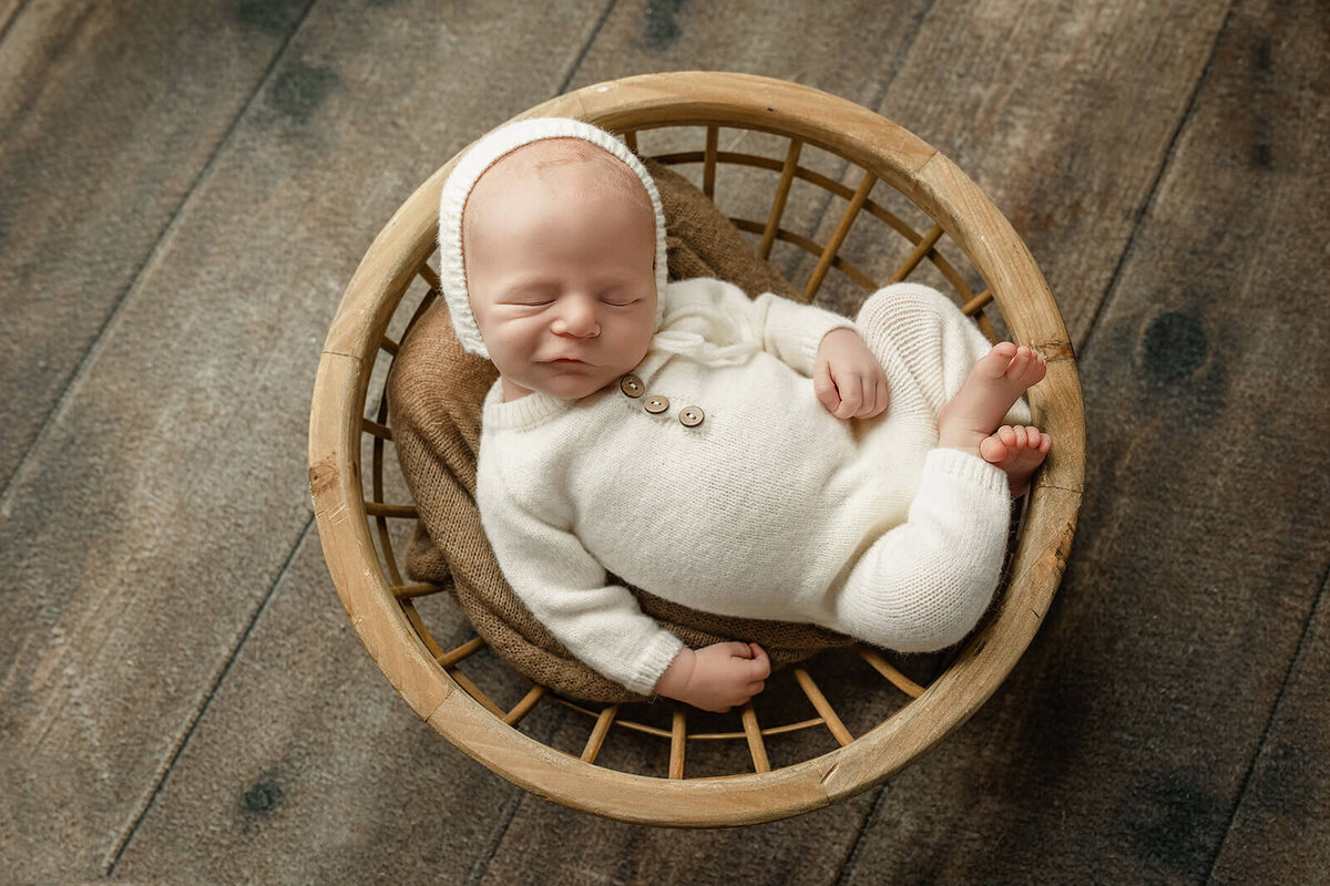 newborn boy in a white romper with matching white bonnet in a round basket with a soft smirk on his face for newborn photos 