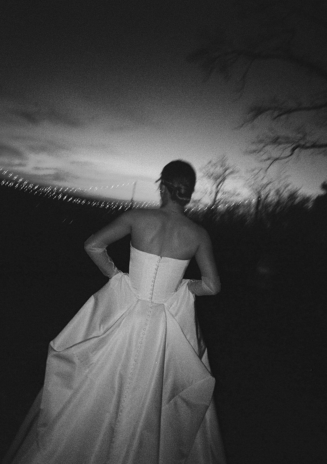 Black and white film photograph of a bride in a strapless ballgown walking outside at Burritt on the Mountain at sunset, with string lights glowing in the distance.