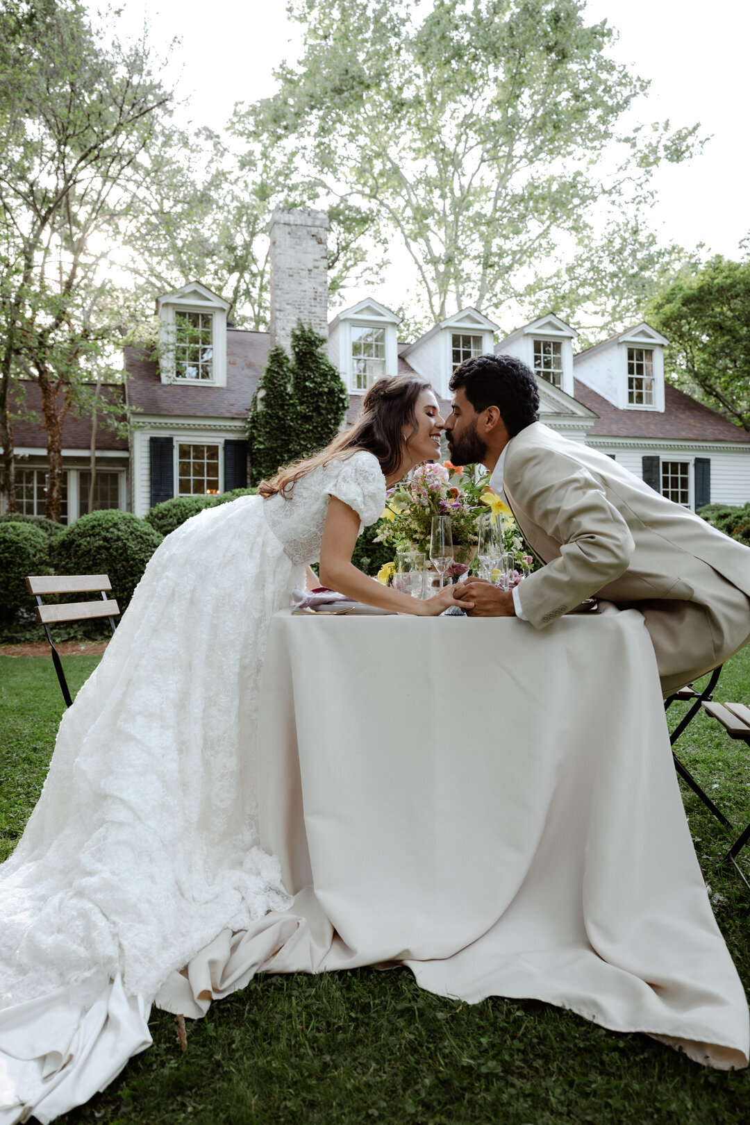 Bride and groom sharing a quiet moment at a table set up in a garden, surrounded by spring greenery.