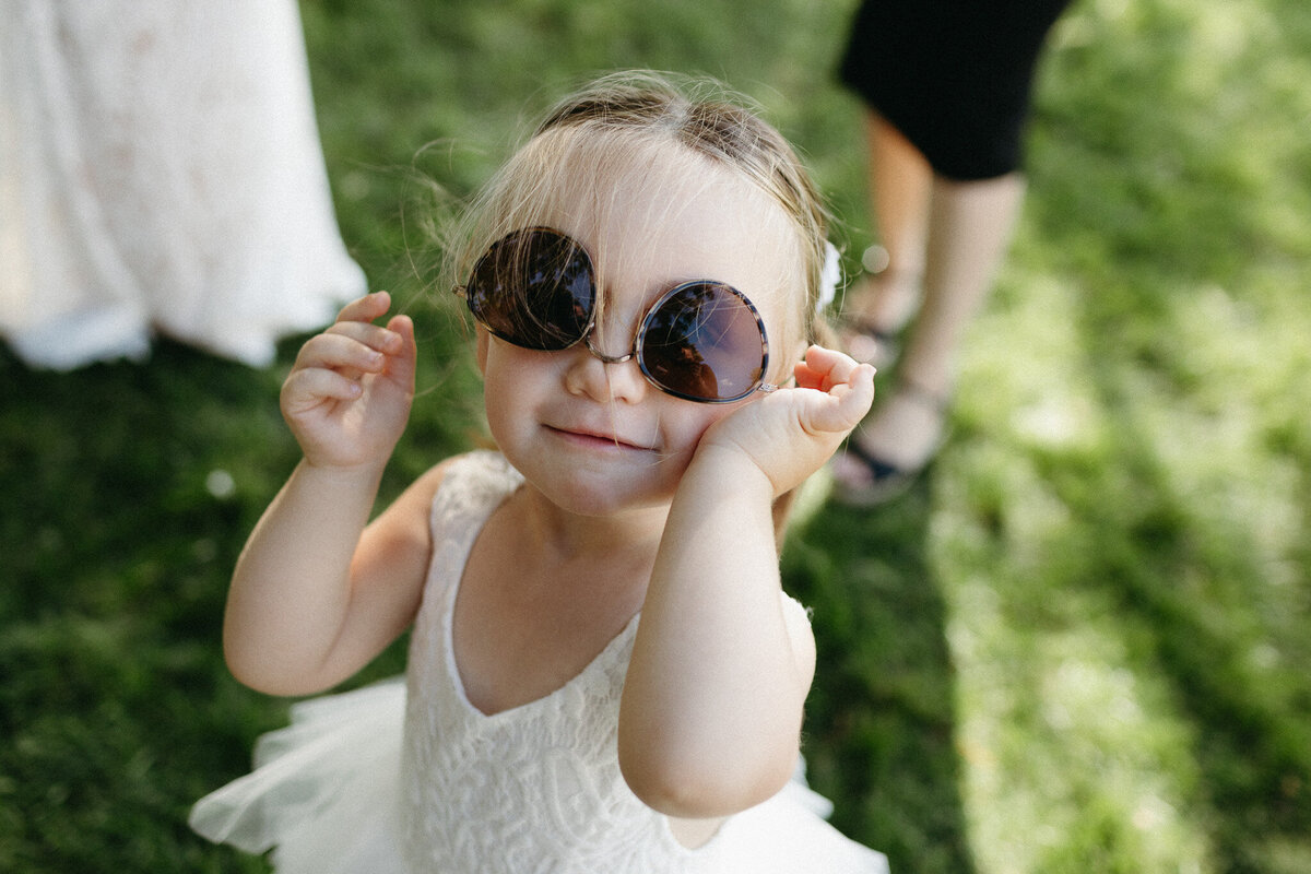 A young child in a white dress smiles joyfully, wearing large, round sunglasses while playing on the grass. Captured with the keen eye of a Toronto photographer, this charming moment epitomizes pure bliss and innocence under the open sky.