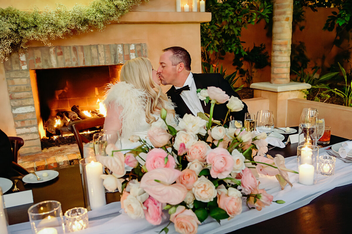 Bride and groom kiss while sitting at their wedding reception head table with beautiful pink and white floral arrangement