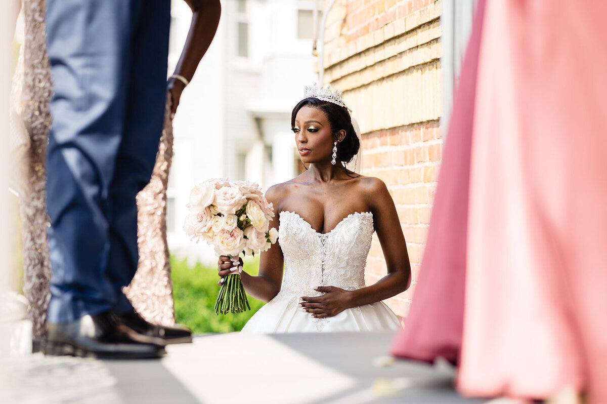 A bride in a strapless wedding dress and tiara holding a bouquet, with a subtle gaze to the side, standing in front of a brick building