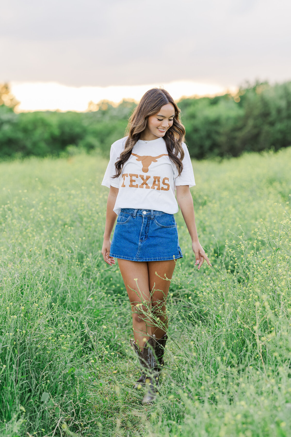 Senior girl walking through tall grass at sunset in a Texas Longhorns T-shirt and denim skirt, captured at Arbor Hills in Plano, Texas.