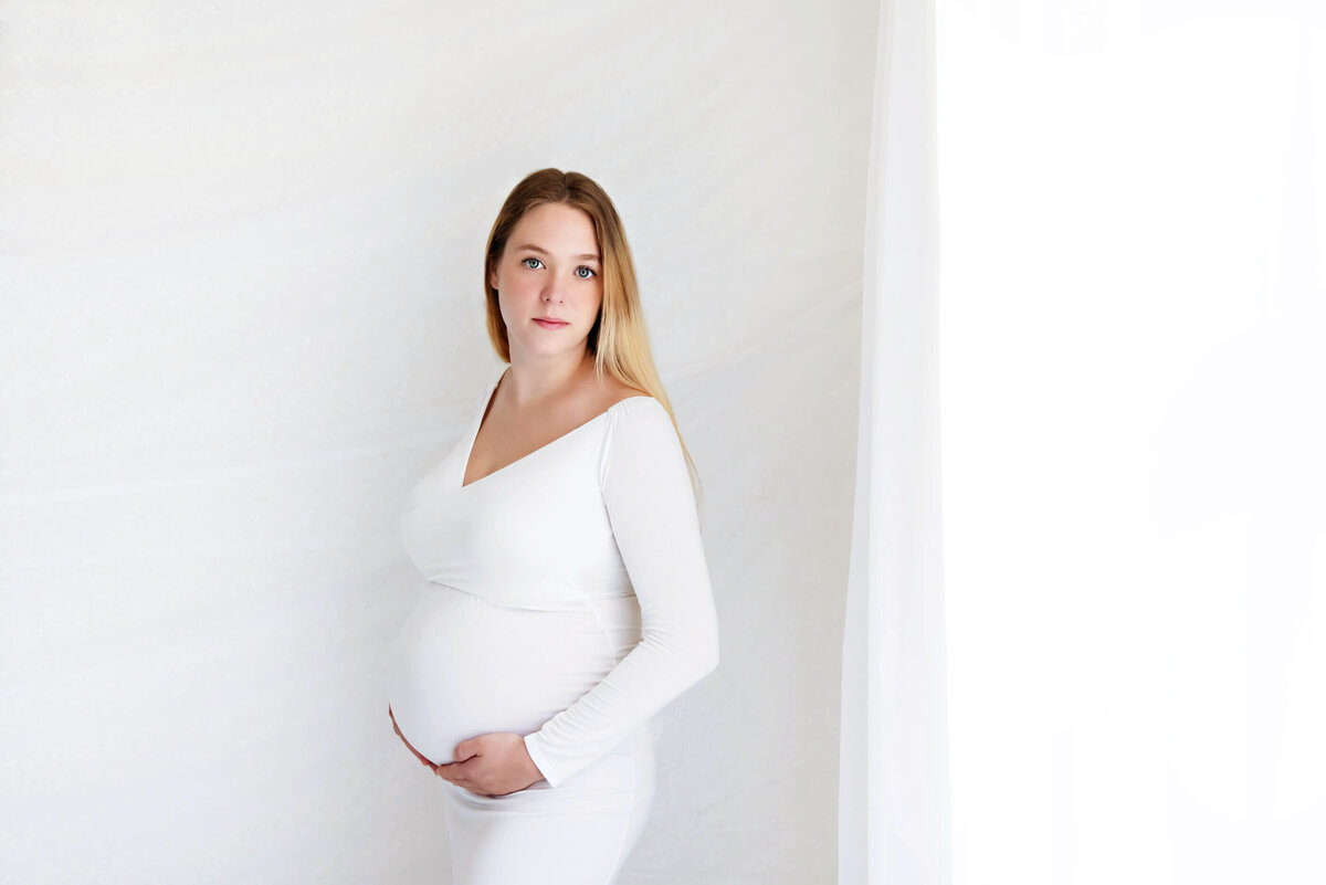 Blonde woman wearing a white maternity dress on a white background for her Burlington studio maternity photography session
