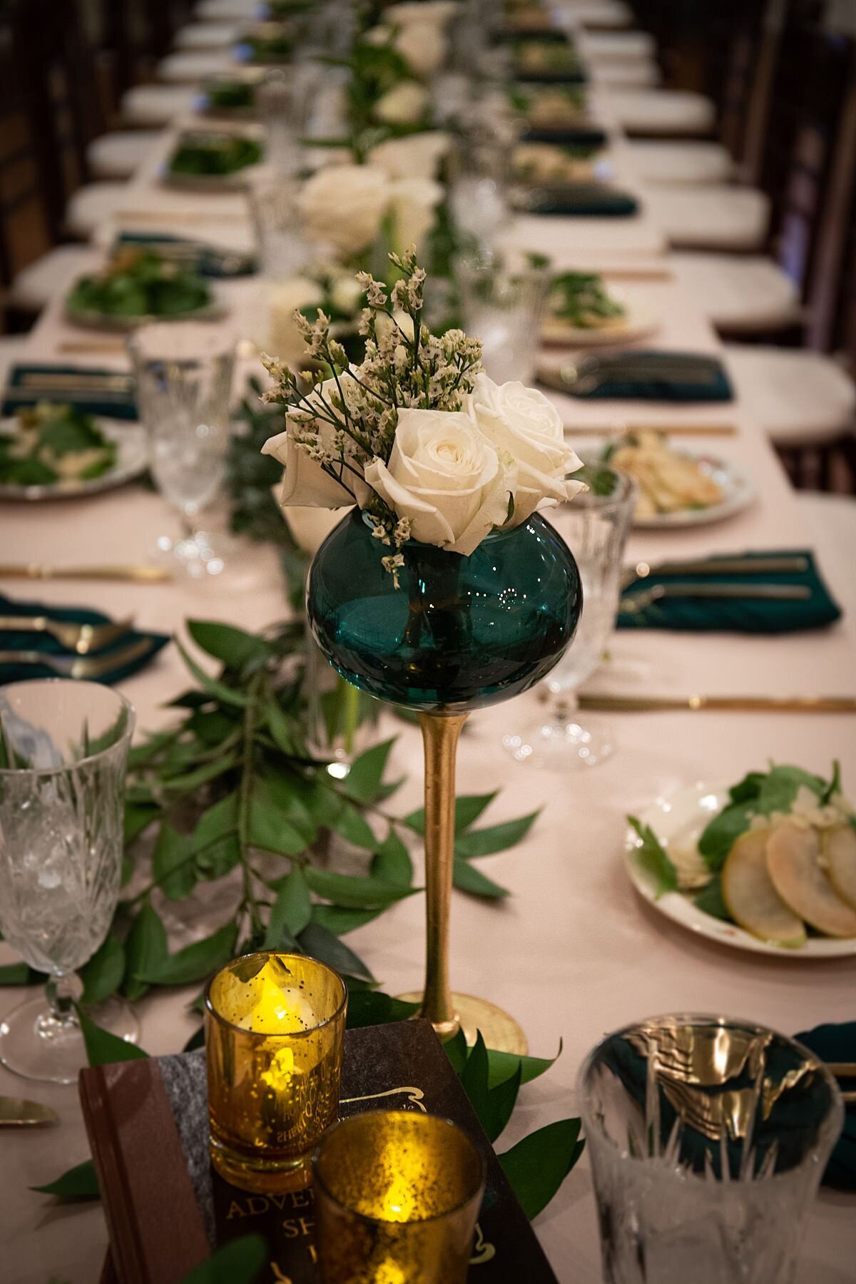 long banquet table with white table cloths are set with emerald green napkins, gold flatware, cut crystal goblets and pear salads. Gold votive candles, emerald green glass vases and long garlands of greenery with white roses run the length of the long table at Oaklands Mansion.