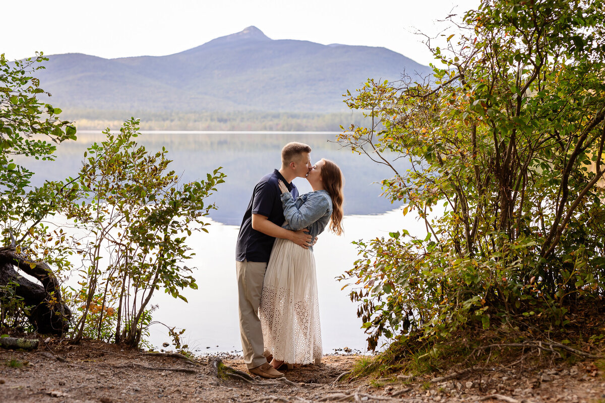Couple kissing at Chocorua Lake framed by beautiful green bushes in North Conway NH Engagement Photography Session