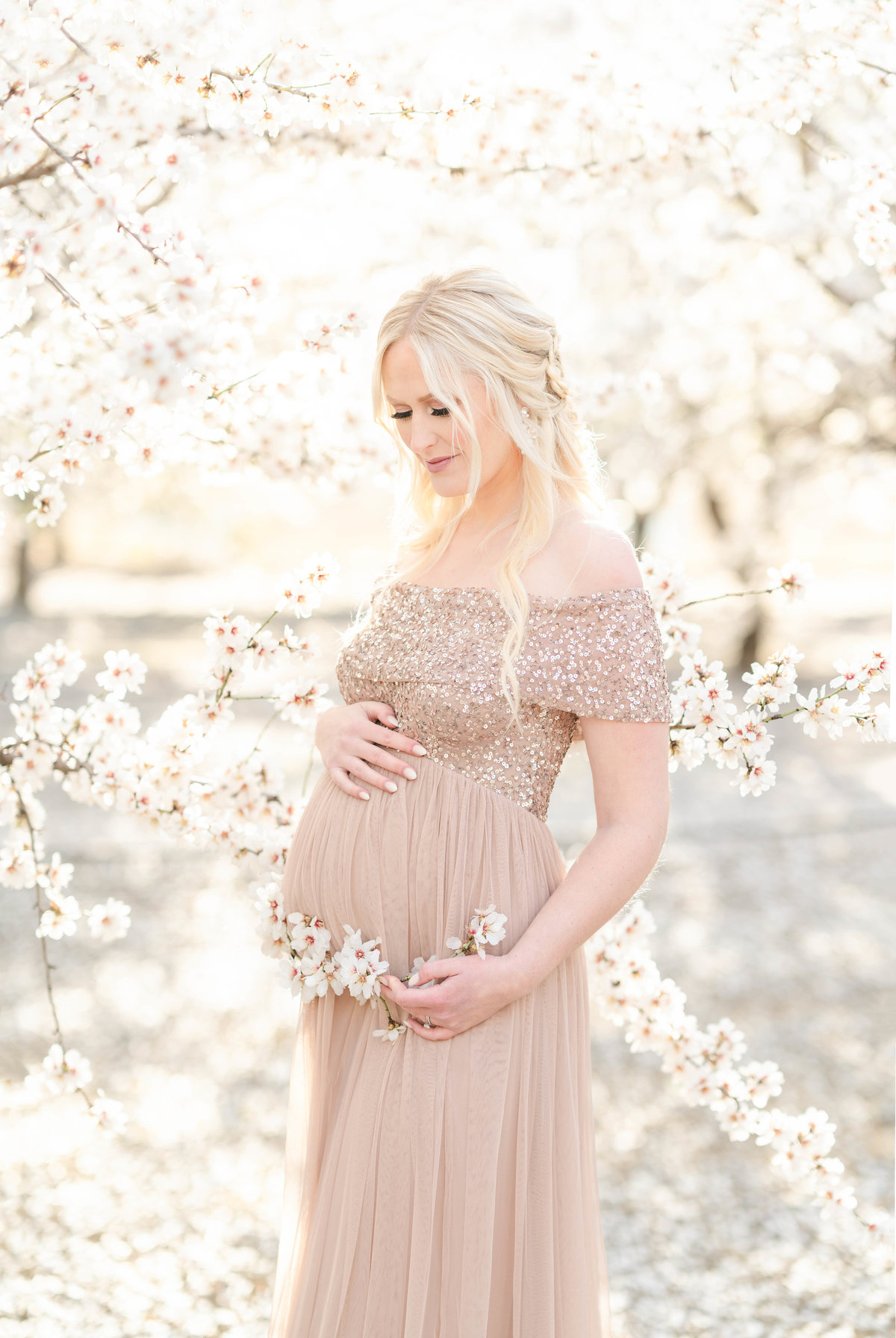 An expecting mother is wearing a dusty rose gown while standing in an almond blossom field caressing her beautiful bump photographed by Bay area photographer, Light Livin Photography.