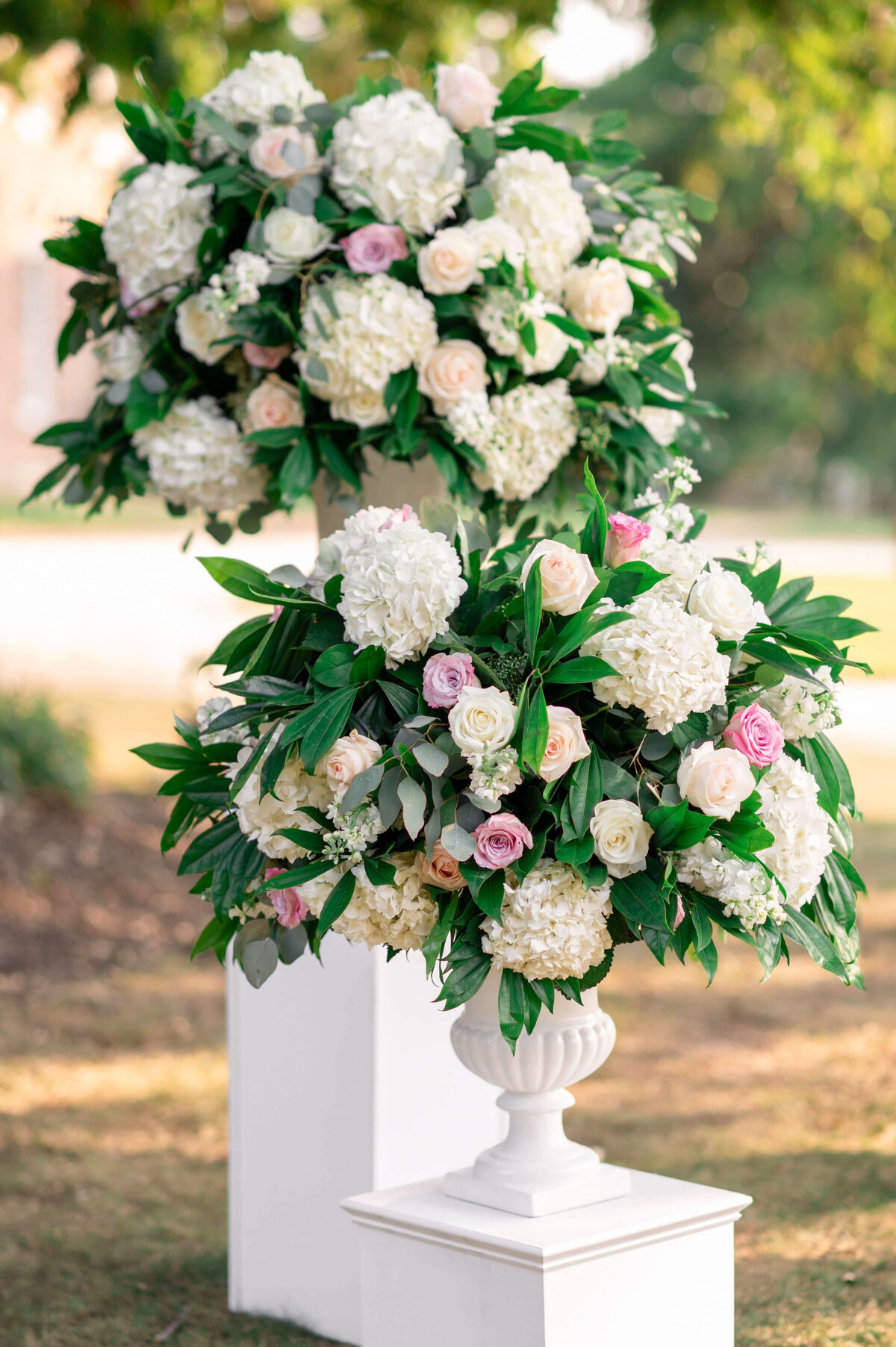 Elegant floral arrangements featuring white hydrangeas and pink roses, displayed in white pedestal vases. Lush green foliage complements the flowers, set outdoors with a blurred background of trees and a building.