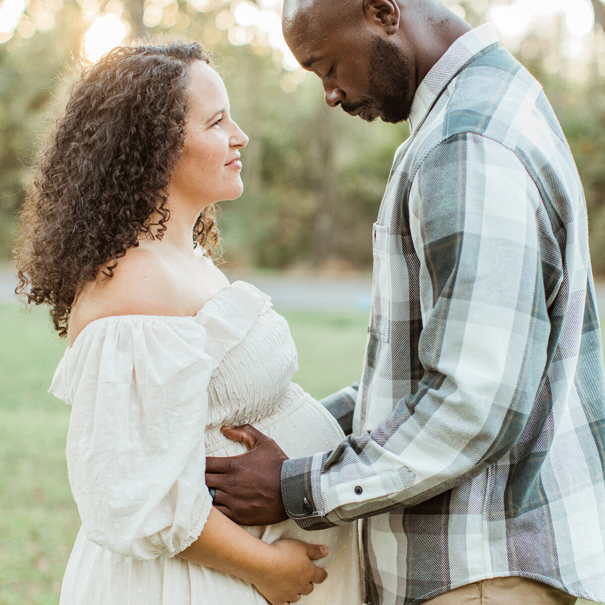 pregnant mom stand across from partner outside as they look down at mom’s growing stomach