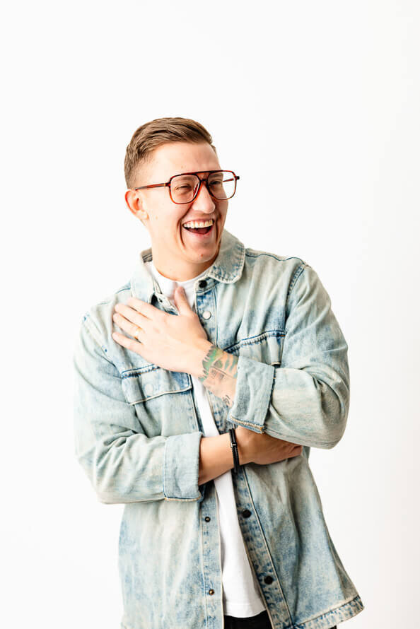 Man in jean jacket on a white background laughs and puts hand on chest.
