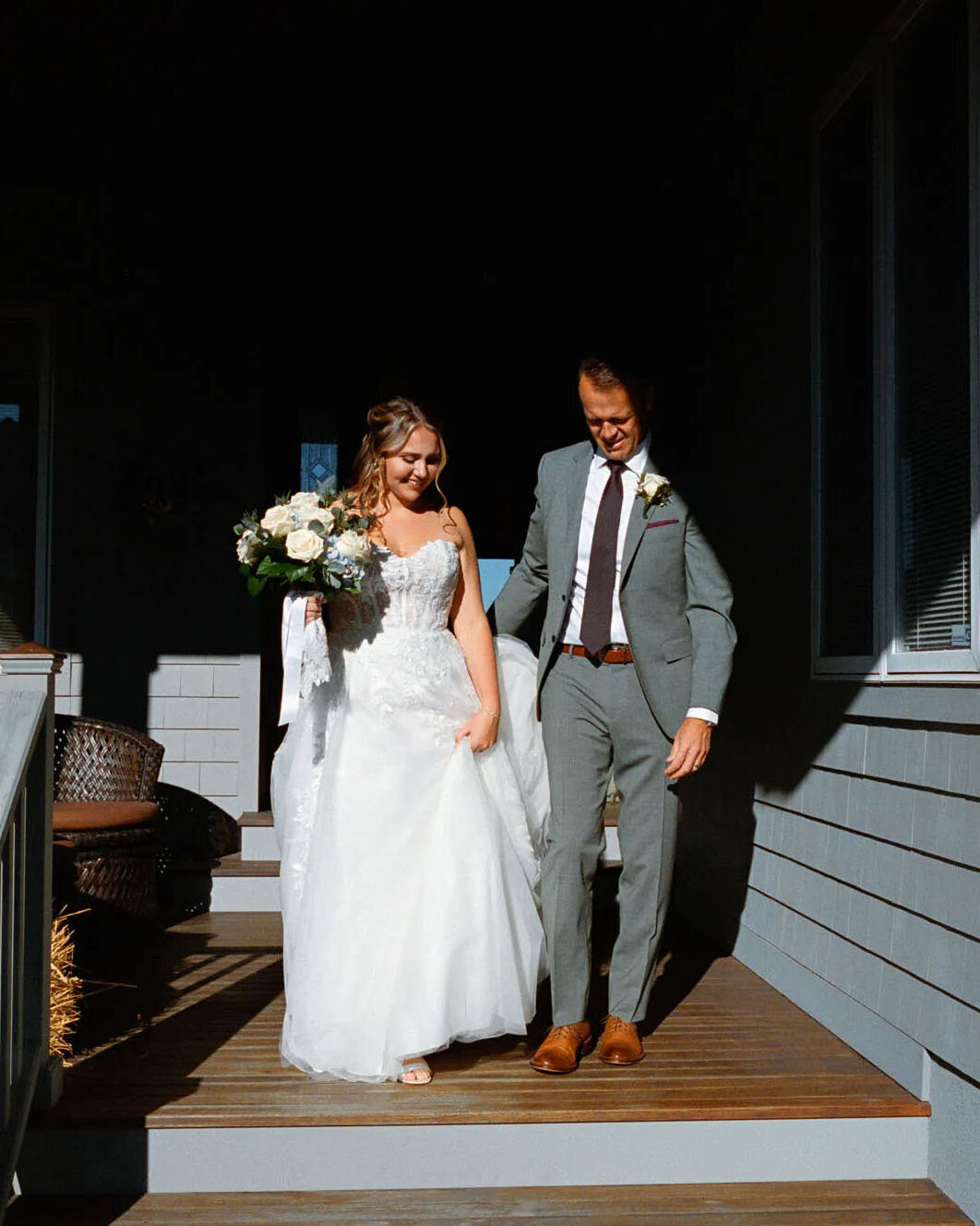 A bride in a white gown holds a bouquet and walks down porch steps beside a man in a gray suit, both smiling, as sunlight casts shadows on a gray house—captured beautifully by a film photographer NJ.